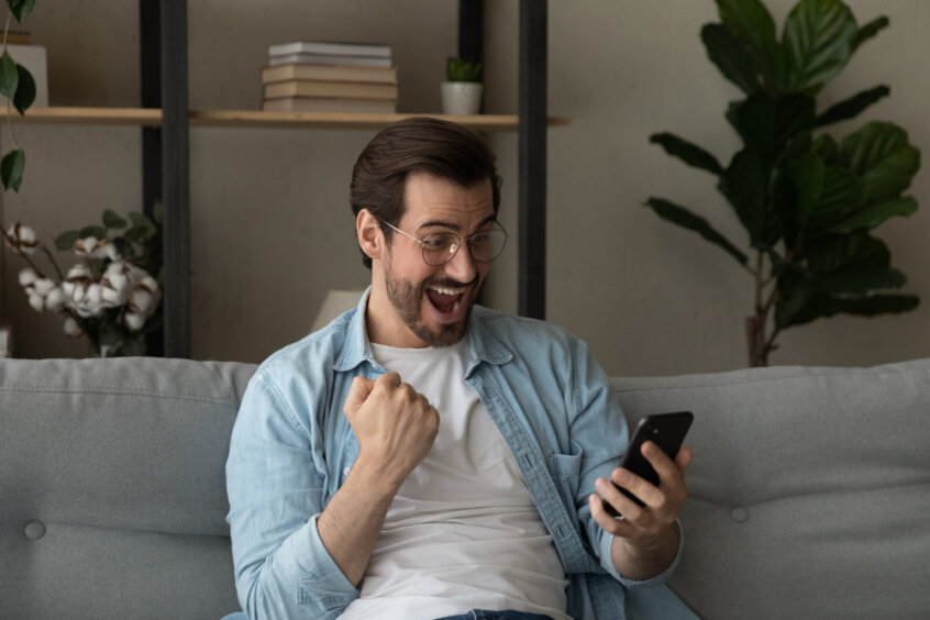 Excited,Overjoyed,Young,Man,Sit,On,Sofa,Holding,Smartphone,Feeling